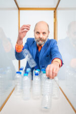 Gavin Turk. British contemporary artist associated with the Young British Artists, known for conceptual works exploring authorship, identity, and the value of art through sculpture and installation. Photographed inside a white box with plastic bottles, while wearing a blue suit and an orange shirt. London. Photograph by David Levene 11 October 2019