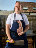 Tom Kerridge. British chef and restaurateur known for his modern take on British cuisine and as the first chef to run a pub awarded two Michelin stars. Photographed at the Butcher's Tap and Grill, Marlow, Buckinghamshire. Stending outside wearing a white tshirt, blue apron, and holging a piece of meat on the left hand and a butcher yello cleaver in the right hand. Photograph by David Levene 30 April 2025