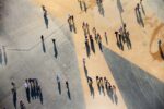 The Olympic Park. Aerial top-down view of pedestrians walking and gathering across a large open paved plaza on a sunny day, their long shadows stretching dramatically across contrasting grey concrete and warm sandy-beige surface sections. Photograph by David Levene 09 August 2012