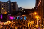 KERB. Peckham. Claude responded: Elevated evening view of a busy outdoor street food market in Peckham, South London, with crowds of people gathered around food stalls and vendors, large illum…Elevated evening view of a busy outdoor street food market in Peckham, South London, with crowds of people gathered around food stalls and vendors, large illuminated "KERB" letters and a neon "Peckham" sign against a brick wall, colourful purple and orange lighting, surrounded by urban residential and commercial buildings at dusk. London. Photograph by David Levene 19 July 2013