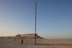 A figure in black standing in a desert landscape beside a tall vertical pole, Doha, Qatar. Photograph by David Levene 30 March 2022