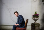 Fraser Nelson, Scottish journalist and editor of The Spectator magazine, seated on a wooden chair against a weathered exterior wall, London. Photograph by David Levene, 3 February 2014.