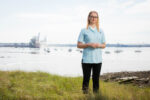 An NHS nurse in uniform standing on a grassy riverbank with moored boats and a port visible in the background, England. Photograph by David Levene, 14 April 2016