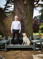 Ken Follett, Welsh author best known for his historical novels including The Pillars of the Earth and the Kingsbridge series, standing barefoot on a garden bench beside a yellow Labrador in front of a large tree, England. Photograph by David Levene, 28 July 2020