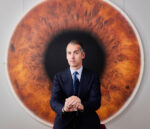 Fabrizio Moretti, Italian Old Master art dealer and founder of Moretti Fine Art, seated in front of a large-scale photorealist painting of a human eye at his London gallery. Photograph by David Levene, 26 May 2022