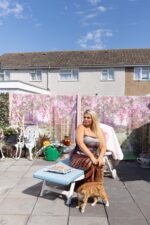 Trewley Precious Temple seated on a sun lounger in a residential garden with her cat, wearing a strapless printed bodycon dress. Photograph by David Levene, 03 August 2026